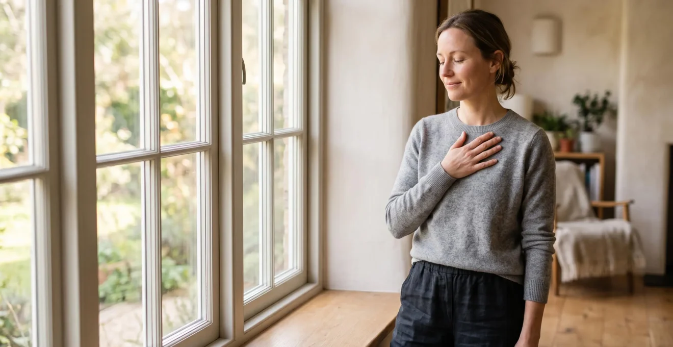 Personne debout près d'une fenêtre lumineuse, main posée sur sa poitrine dans un geste d'auto-compassion, symbolisant une relation apaisée avec son corps